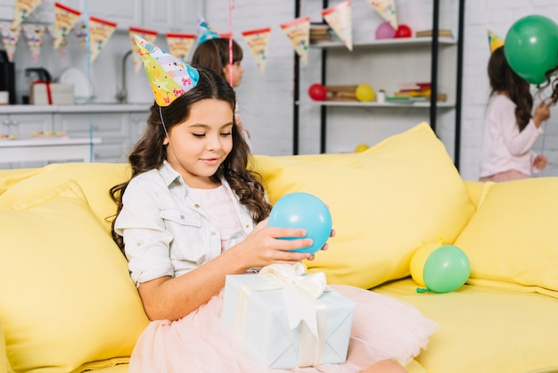 Ragazza graziosa di compleanno che si siede sul pallone della tenuta del sofà a disposizione che esamina il contenitore di regalo