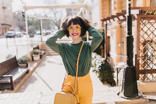 Ragazza graziosa di risata che gioca con i suoi capelli castani corti. Ritratto all'aperto di splendida donna bianca in abiti casual.