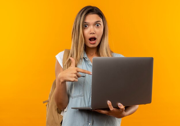 Ragazza graziosa giovane impressionata dell'allievo che porta la tenuta posteriore della borsa e che indica al computer portatile isolato sull'arancia