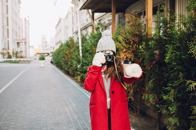 Ragazza graziosa in cappotto rosso e cappello lavorato a maglia che cammina sulla strada. Fa foto del caffè in mano.