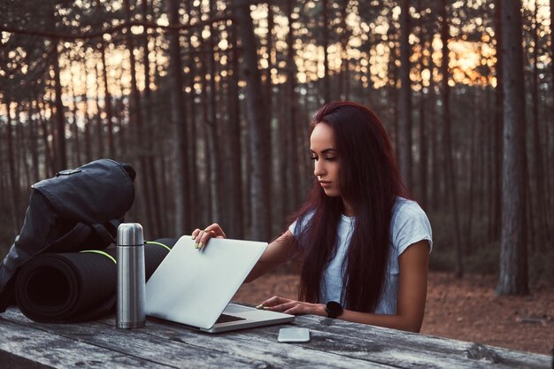 Ragazza hipster che apre un computer portatile per lavorare su una panca di legno mentre si fa una pausa in una bellissima foresta autunnale.