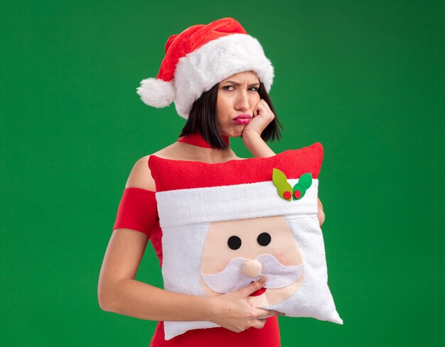 Ragazza imbronciata che porta il cappello della santa che tiene il cuscino del babbo natale che tiene la mano sul mento isolata sulla parete verde