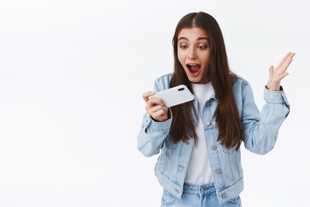 Ragazza impressionata eccitata ed elettrizzata che grida allo smartphone stupita alzando la mano trionfante tenendo il telefono cellulare orizzontalmente fissare lo schermo stupito sfondo bianco Concetto di tecnologia