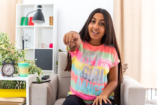 Ragazza in abiti casual che punta con il dito indice sorridendo allegramente felice e positiva seduta sulla sedia in un soggiorno luminoso