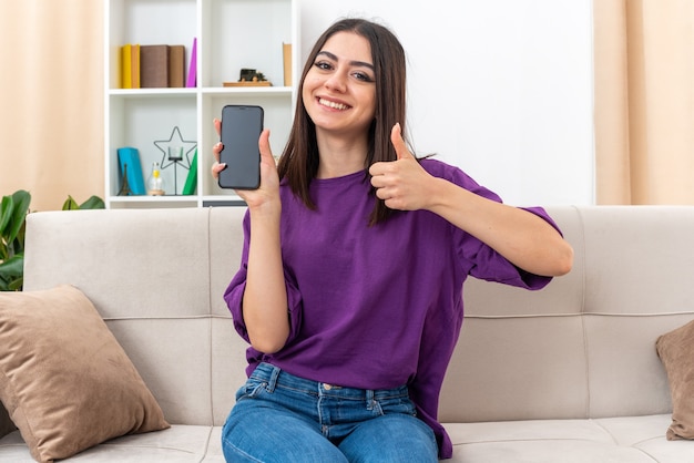 Ragazza in abiti casual che tiene in mano uno smartphone che sorride allegramente mostrando i pollici seduta su un divano in un soggiorno luminoso