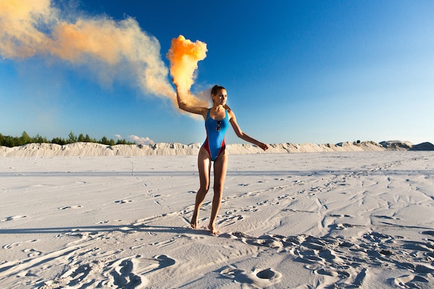 Ragazza in blu swim-suit danze con il fumo arancione sulla spiaggia bianca