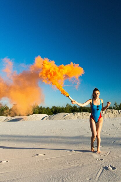 Ragazza in blu swim-suit danze con il fumo arancione sulla spiaggia bianca