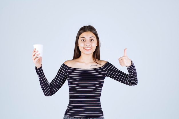 Ragazza in camicia a righe che tiene una tazza di caffè di plastica e che indica un buon gusto.