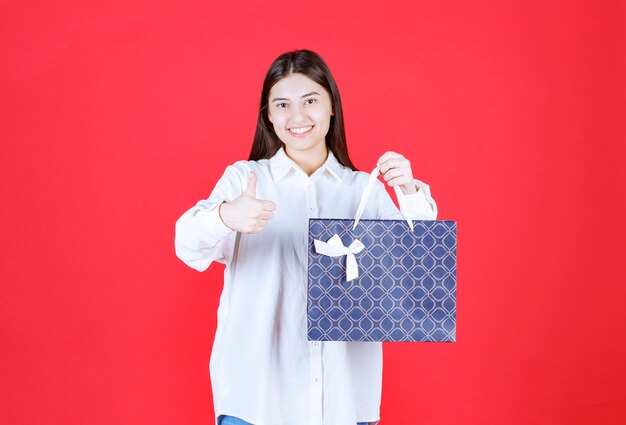 Ragazza in camicia bianca che tiene in mano una borsa della spesa blu e mostra un segno positivo con la mano