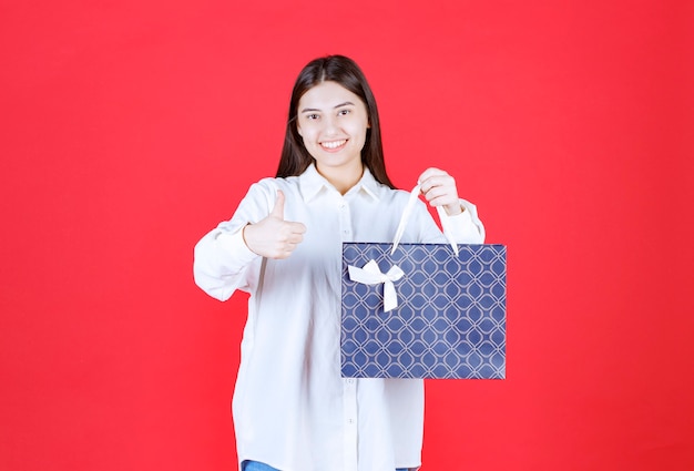 Ragazza in camicia bianca che tiene in mano una borsa della spesa blu e mostra un segno positivo con la mano