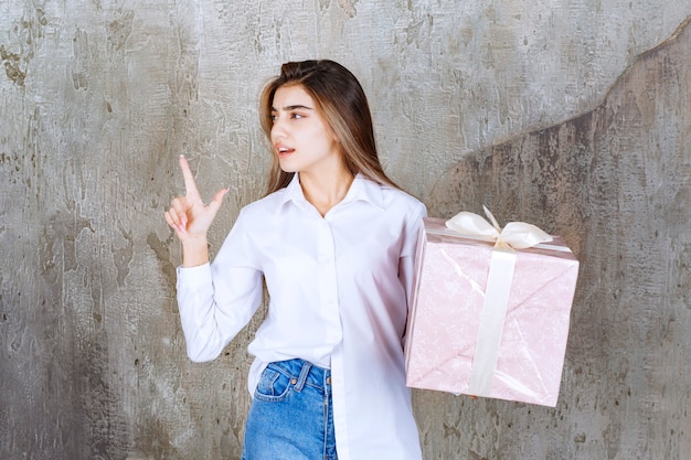 Ragazza in camicia bianca che tiene una scatola regalo rosa avvolta con un nastro bianco e che ha una buona idea.