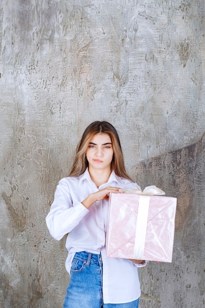Ragazza in camicia bianca con in mano una confezione regalo rosa avvolta con un nastro bianco e sembra confusa ed esitante.