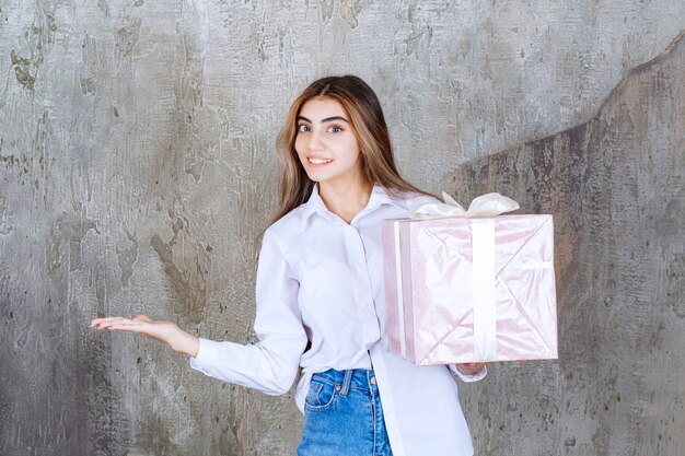 Ragazza in camicia bianca con in mano una scatola regalo rosa avvolta con un nastro bianco, notando il suo partner e chiedendogli di venire a riceverlo