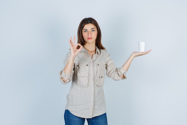 ragazza in camicia con in mano una tazza di caffè in plastica, che mostra il segno ok e sembra soddisfatta.