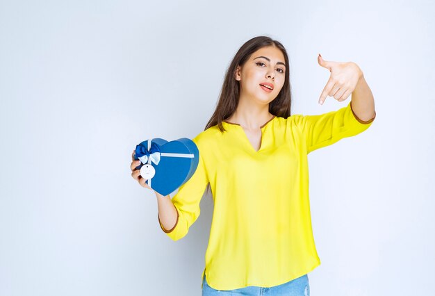 Ragazza in camicia gialla che tiene e promuove una confezione regalo a forma di cuore blu.