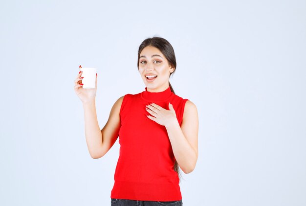 Ragazza in camicia rossa che tiene una tazza di caffè bianca e che indica se stessa.