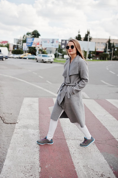 Ragazza in cappotto grigio con occhiali da sole e borsetta che cammina sul passaggio pedonale