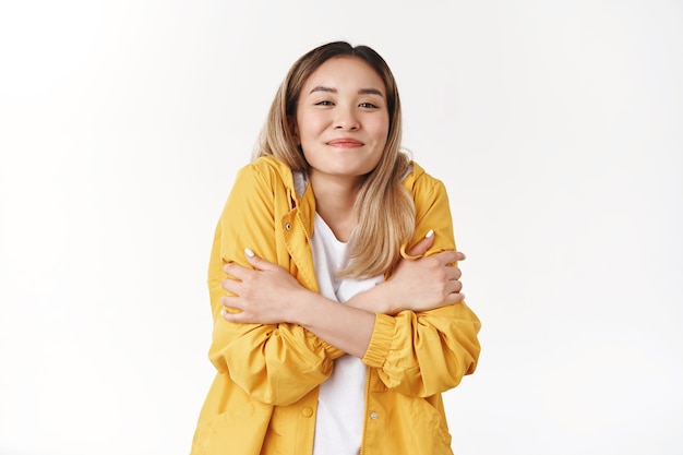 Ragazza in fase di riscaldamento indossare giacca gialla sensazione di freddo a piedi la sera estate vacanza al mare abbracciandosi stretto sorridente felice guardare soddisfatto allegro in piedi muro bianco contento