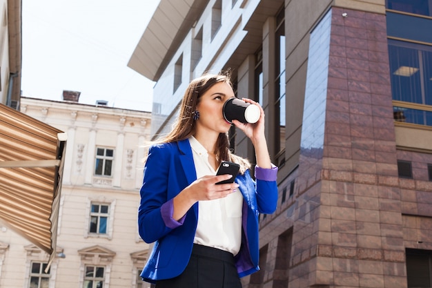 Ragazza in giacca blu brillante si trova con smartphone e caffè sulla strada