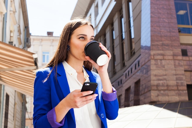 Ragazza in giacca blu brillante si trova con smartphone e caffè sulla strada