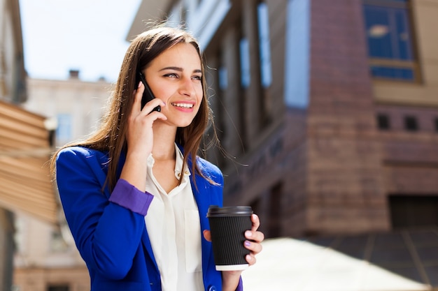 Ragazza in giacca blu brillante si trova con smartphone e caffè sulla strada