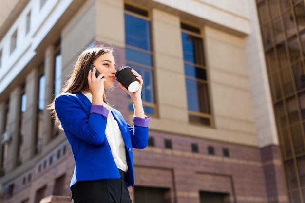 Ragazza in giacca blu brillante si trova con smartphone e caffè sulla strada