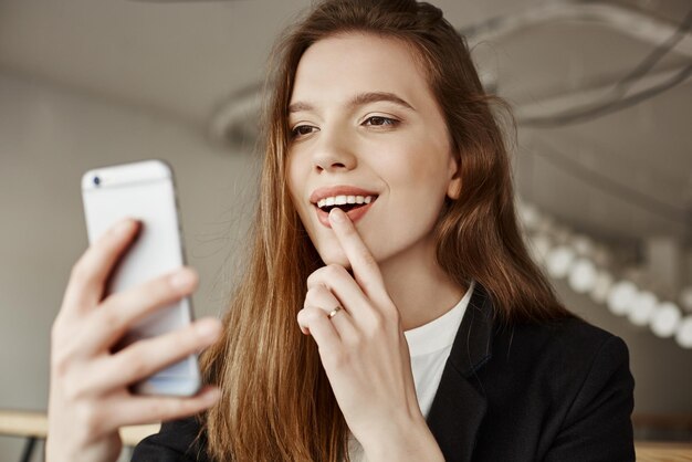 Ragazza in posa per selfie Scatto al coperto di bella donna caucasica seduta in un bar con il dito indice sul labbro per dare un aspetto sensuale sorridente allo schermo mentre si scatta una foto sullo smartphone