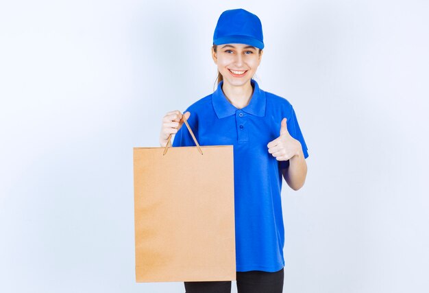 Ragazza in uniforme blu che tiene un sacchetto della spesa del cartone e che mostra il segno di soddisfazione.