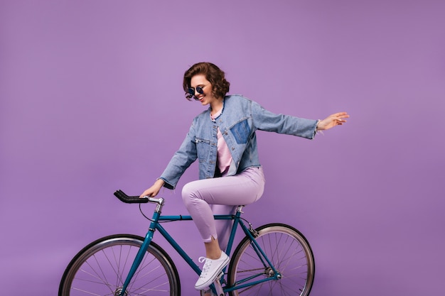 Ragazza incantevole sorridente che si siede sulla bicicletta blu. modello femminile di buon umore