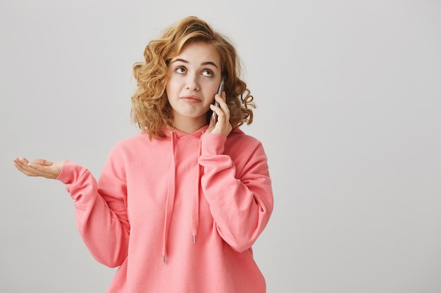 Ragazza indecisa all'oscuro che parla al telefono, scrollando le spalle perplessa