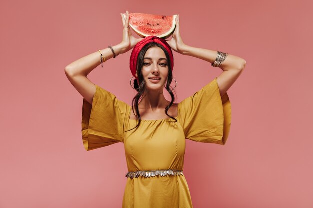 Ragazza indiana con capelli ondulati neri in fascia e vestiti alla moda gialli che tengono l'anguria sulla sua testa sulla parete rosa