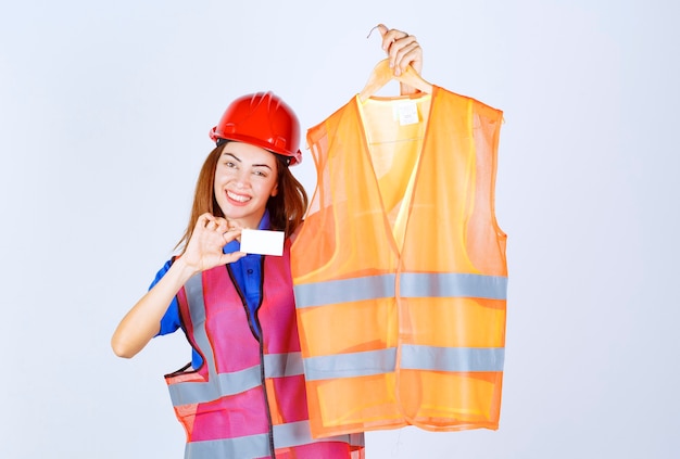 Ragazza ingegnere in uniforme che presenta un pezzo di giubbotto di sicurezza al collega e presenta il suo biglietto da visita.