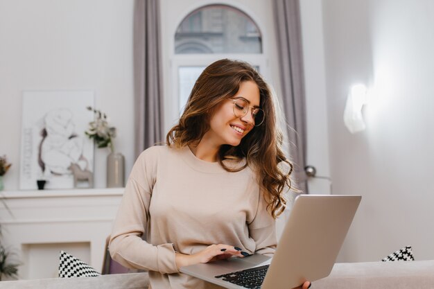 Ragazza intelligente spensierata in camicia beige in posa con un sorriso romantico, lavorando con il computer