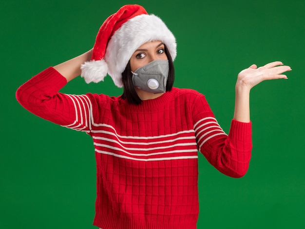 Ragazza interessata che porta il cappello della Santa e la maschera protettiva che guarda l'obbiettivo che tiene la mano sulla testa che mostra la mano vuota isolata su fondo verde