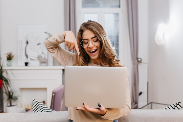 Ragazza interessata con manicure alla moda che scherza a casa durante il servizio fotografico con il laptop