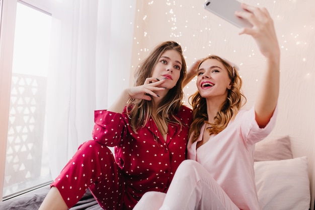 Ragazza interessata in abito da notte rosso che guarda al telefono di un amico mentre le scatta una foto. Felice donna bionda in pigiama rosa utilizza lo smartphone per selfie.