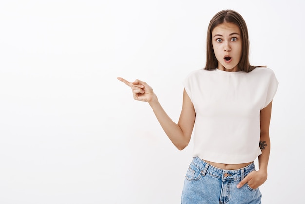 Ragazza interrogata e scioccata che indica la bocca aperta a sinistra per la sorpresa e lo stupore tenendo la mano in tasca in piedi e iniziando a tremare dalla scena incredibile