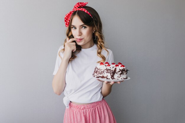 Ragazza ispirata con capelli ondulati degustazione torta di compleanno. incantevole modello femminile con torta in posa.