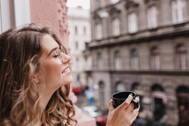 Ragazza ispirata con occhiali alla moda in posa con gli occhi chiusi su sfondo sfocato della città durante l'ora del tè. Foto ravvicinata di una sensuale signora dai capelli scuri che si rilassa al mattino con un gustoso caffè.