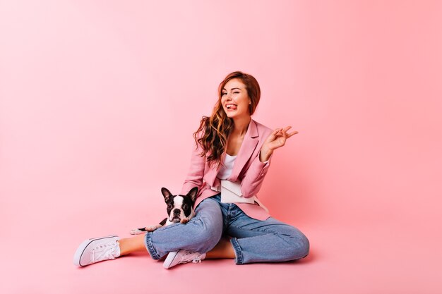 Ragazza ispirata in jeans e scarpe da ginnastica in posa con il bulldog. Ritratto dell'interno del modello femminile dello zenzero entusiasta che si siede sul pavimento con il cucciolo.