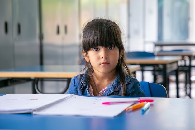 Ragazza latina seria che si siede al banco di scuola e che guarda davanti