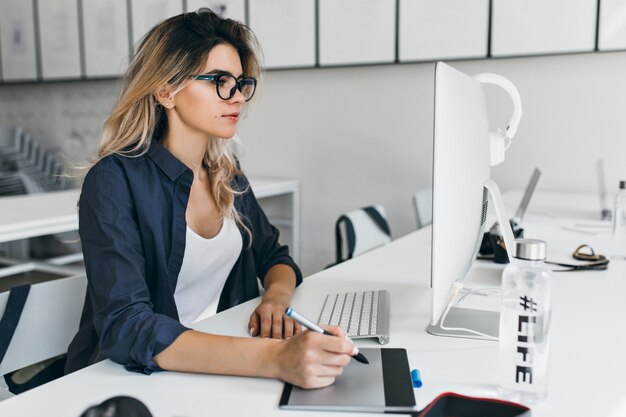 Ragazza leggermente abbronzata in occhiali e camicia nera che lavora con il computer in un ufficio accogliente