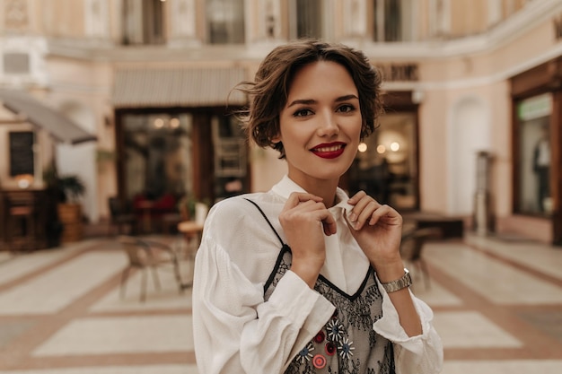 Ragazza meravigliosa con capelli corti e labbra luminose che guardano nella fotocamera in strada Donna bruna in camicia bianca con pizzo nero sorridente in città