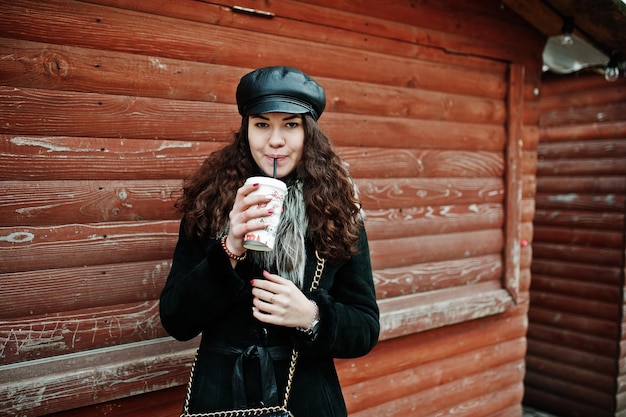 Ragazza messicana riccia con cappuccio in pelle e tazza di caffè in plastica a portata di mano contro il mercato in legno