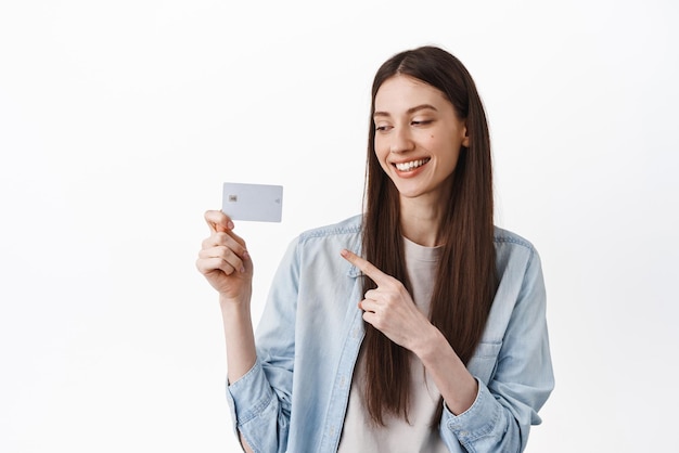 Ragazza moderna che sorride indicando e guardando la carta di credito che consiglia il pagamento senza contatto bancario durante la pandemia in piedi su sfondo bianco