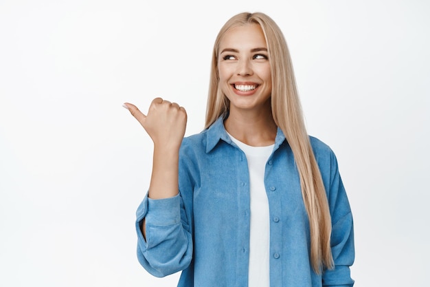 Ragazza moderna felice che punta e guarda a sinistra con un sorriso soddisfatto che consiglia negozio che mostra banner di vendita di pubblicità del negozio in piedi su sfondo bianco