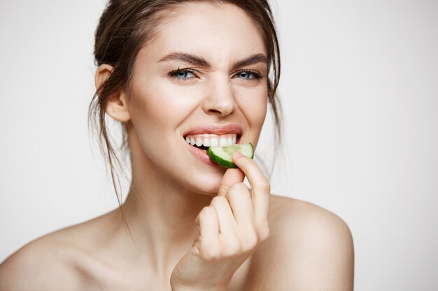 Ragazza naturale abbastanza giovane con pelle pulita perfetta che esamina macchina fotografica che mangia la fetta del cetriolo sopra fondo bianco. Trattamento facciale.