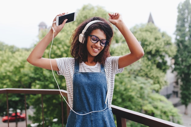 Ragazza nera ispirata con acconciatura riccia alla moda che balla sul balcone con alberi