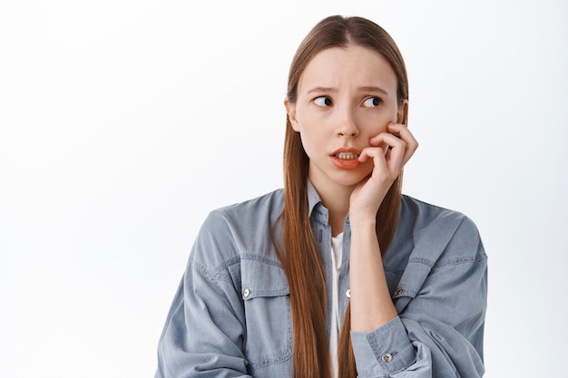 Ragazza nervosa che morde le dita e guarda da parte con un'espressione del viso preoccupata e ansiosa, spaventata da qualcosa, in piedi contro il muro bianco