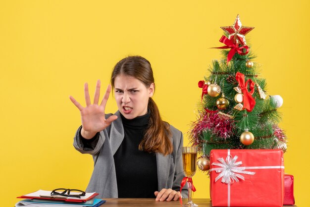 Ragazza nervosa di vista frontale che si siede al tavolo che fa gesto di arresto vicino all'albero di Natale e cocktail dei regali